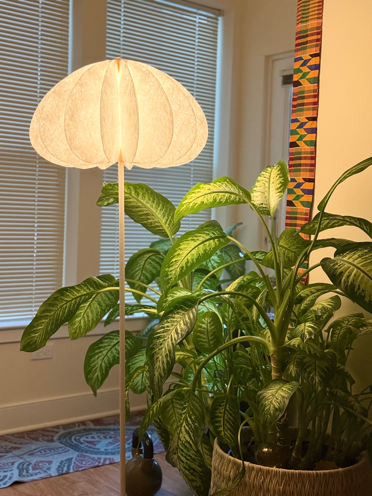 Dandelight floor lamp lit beside a large tropical houseplant near a window