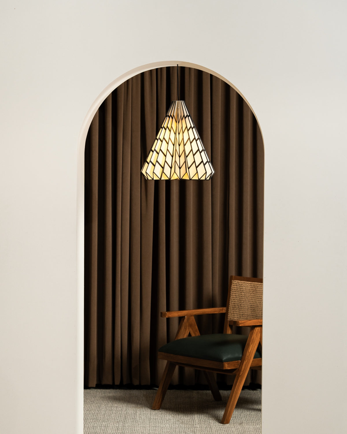 Faceted paper pendant lamp glowing in an arched doorway, against a brown curtain backdrop, above a wood-and-cane armchair